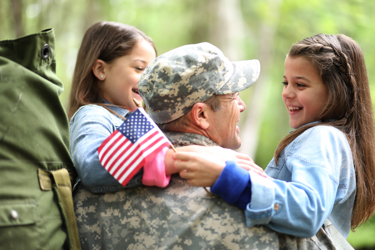 Father in his fatigues uniform holds his two daughters in his arms, with his rucksack still on his back. One of the daughters holds a small American flag.