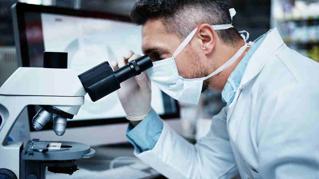 Shot of a mature male with brown hair, mask, gloves, and lab coat using a microscope while conducting medical research for two drugs made by astrazeneca.