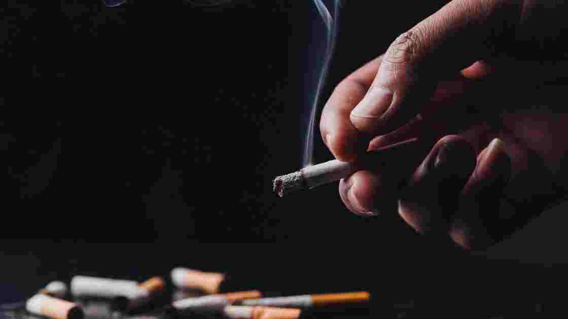Close up of a hand holding a crumpled, smoldering cigarette with smoke above other tobacco cigarettes