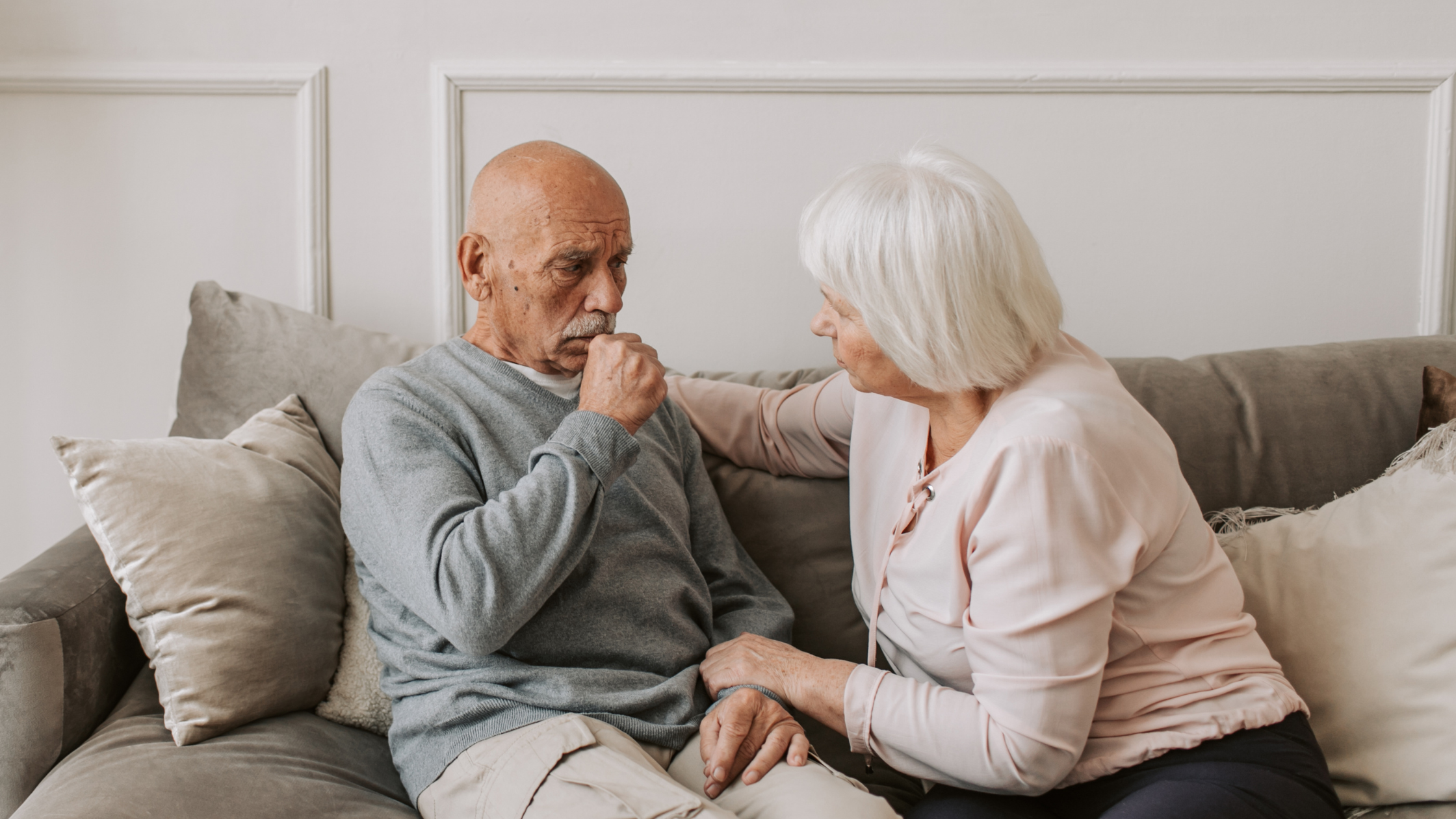 Older bald man sitting on couch with older woman. He is coughing and holding his hand up. Learn the top 5 signs of mesothelioma.