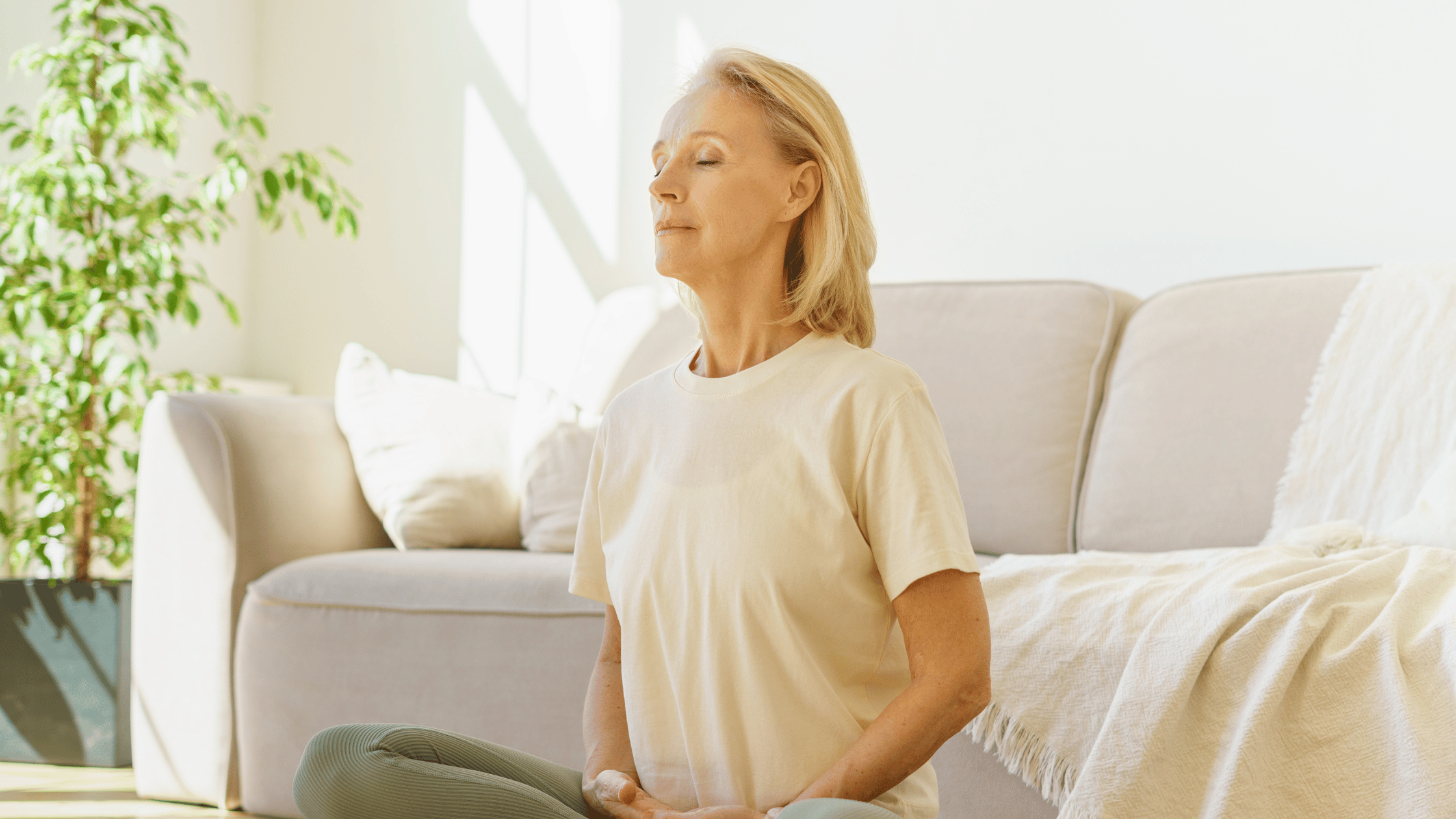 White blonde mature woman sitting on floor with couch and plant in the background practicing breathing exercises for lung health